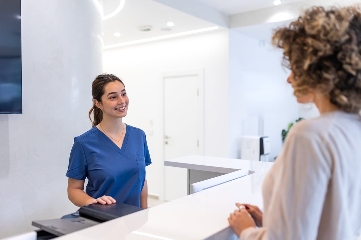 Friendly Medical Receptionist Welcoming Patient at Hospital Front Desk. Smiling Professional in Blue Scrub Uniform at Modern Clinic Reception Area, Healthcare Service and Consultation Concept.